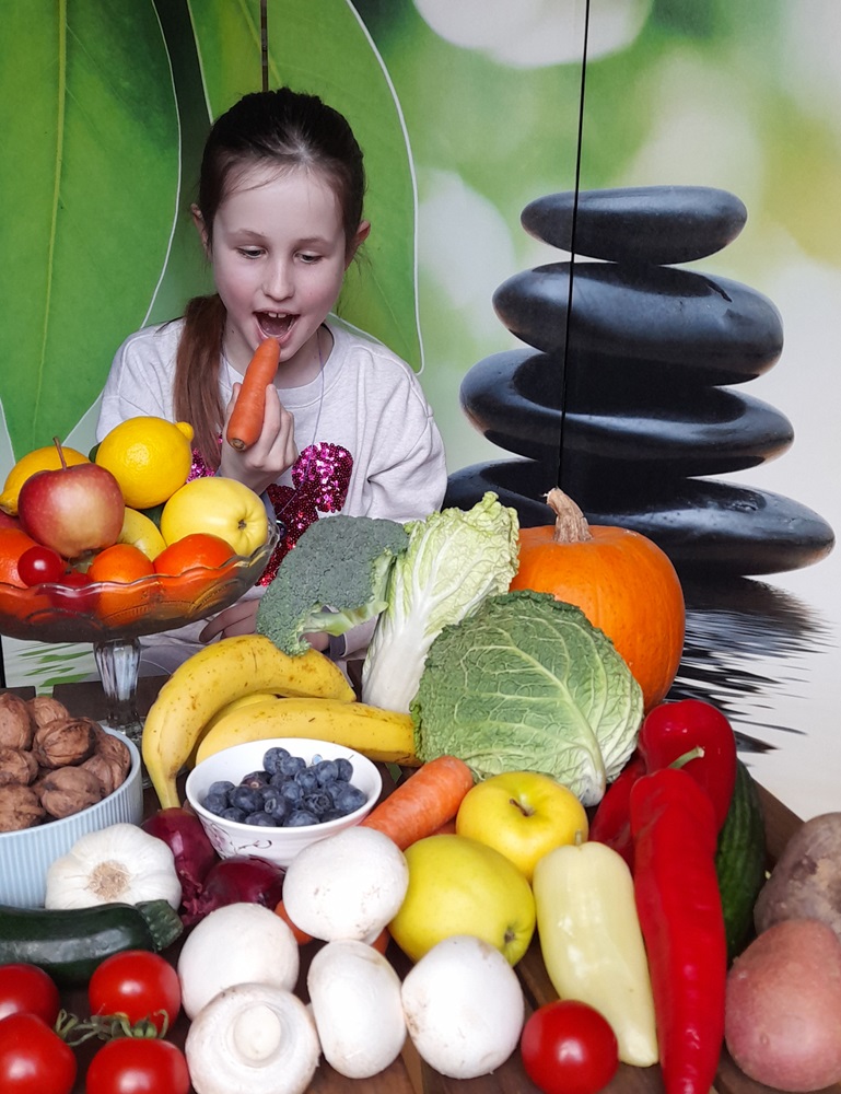 Usmiate dievčatko obklopené zeleninou a ovocím ochutnáva mrkvu